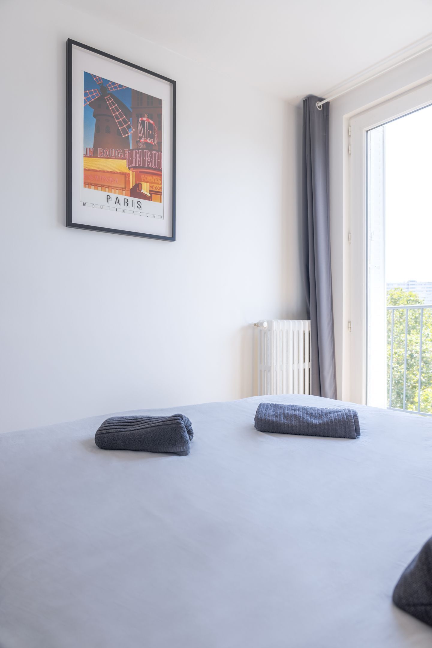 A hotel room with a bed, a radiator, a window with curtains, and a framed poster on the wall.