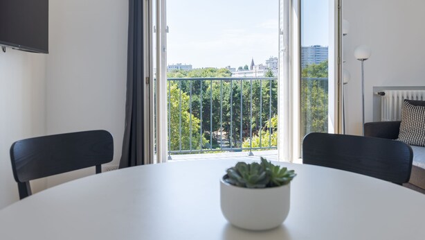 Une chambre moderne avec une table ronde, deux chaises, une plante en pot, un téléviseur et un balcon offrant une vue sur les arbres et les bâtiments.