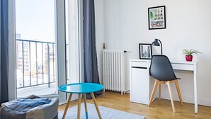 Un appartement moderne avec un plancher en bois, une table ronde bleue, un fauteuil poire gris, un bureau blanc avec une chaise noire, un radiateur et une vue sur la ville à travers une fenêtre.