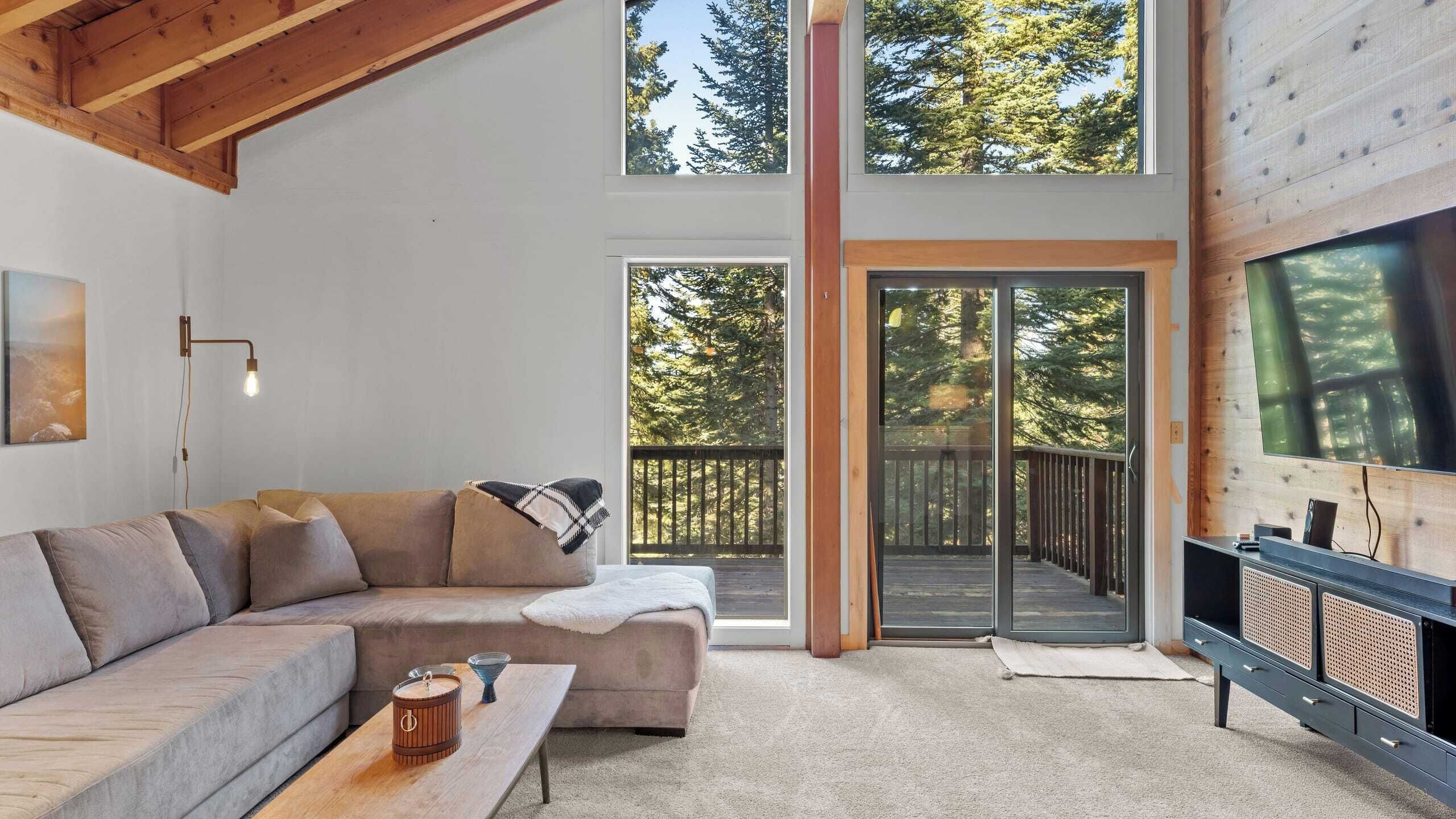 A modern living room with a large sofa, a coffee table, and a TV. There are large windows and a sliding glass door leading to an outdoor deck.