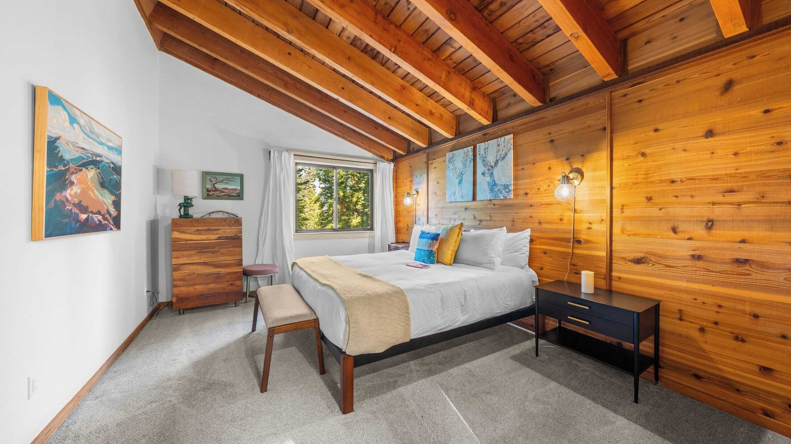 A bedroom with a wooden ceiling, a bed with pillows, a nightstand, a dresser, and a window with a view of trees.