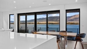 Ein moderner Essbereich mit großem Tisch und Stühlen, große Fenster mit Blick auf einen See und Berge.