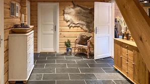 Interieur van een blokhut met een stenen vloer, een bont tapijt, een rieten stoel en een kast.