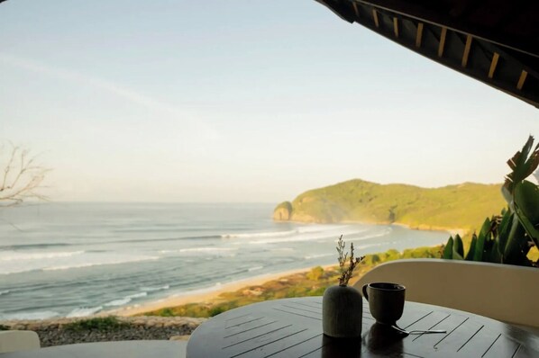 A table with a vase and a cup, overlooking a beach with waves and a distant hill.