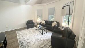 A spacious living room with a large black leather sofa, a coffee table, and a black armchair. There are two windows with blinds, a ceiling fan, and a floor lamp. - High End Downtown 2-Bed Loft, Pet & Child Friendly (Roanoke)