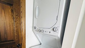 A compact, white front-loading washer and dryer unit with a control panel, positioned in a corner with a wooden door and tiled flooring.