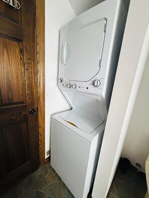 A compact, white front-loading washer and dryer unit with a control panel, positioned in a corner with a wooden door and tiled flooring. - St. Lawrence River home with beach & winter glamping igloo. (Ogdensburg)