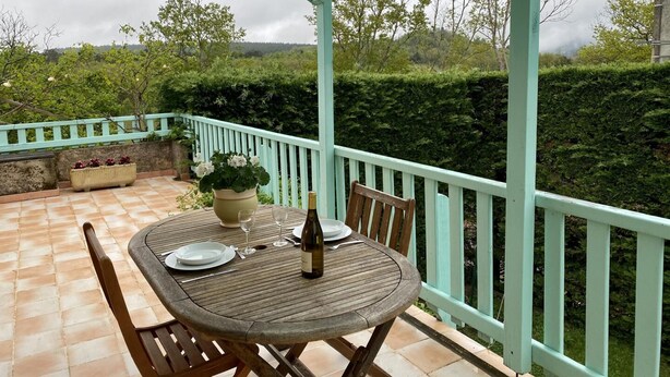 Une terrasse extérieure couverte, avec une table dressée pour deux personnes, une plante en pot et une bouteille de vin.