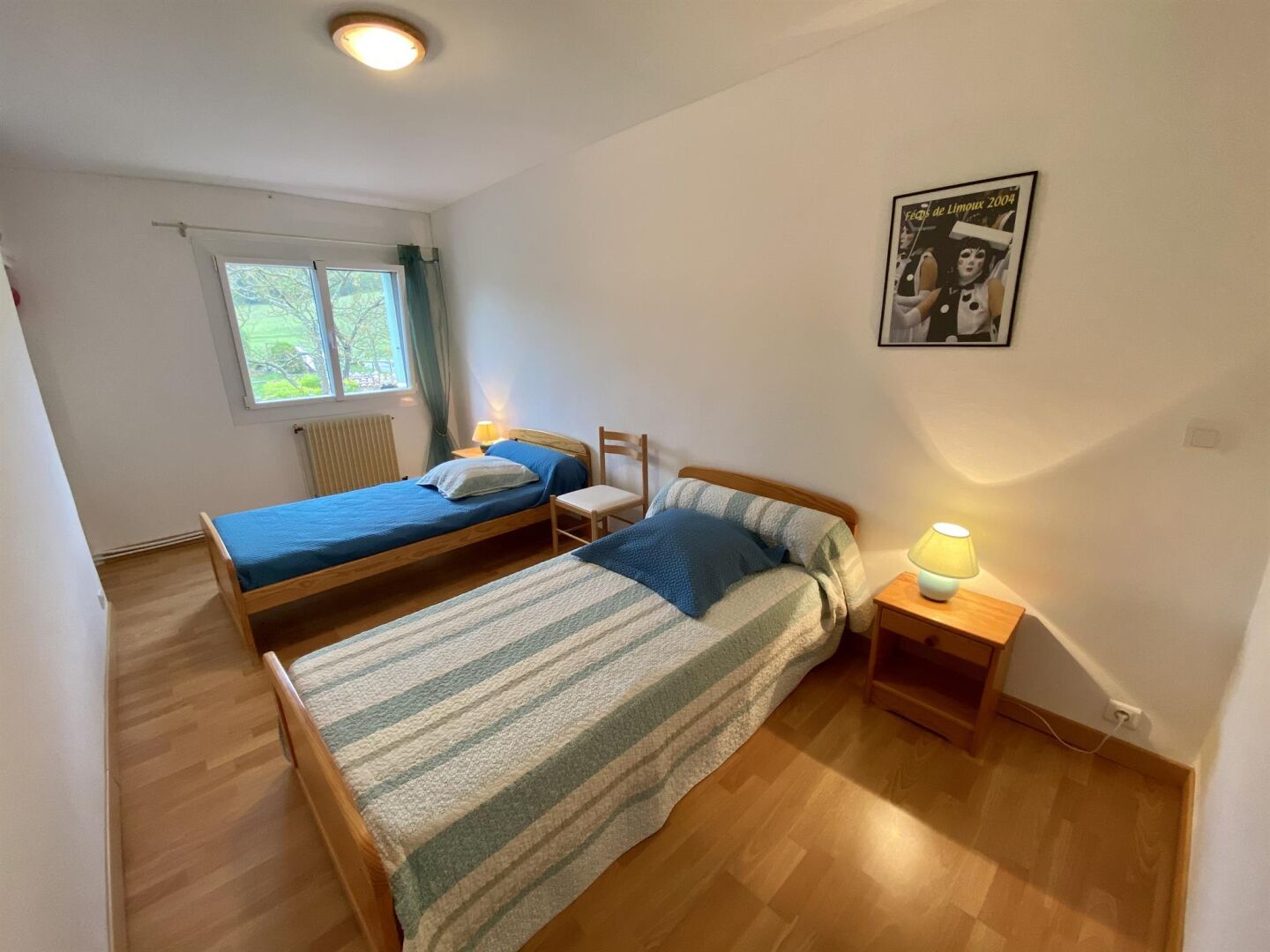 A bedroom with two beds, a window, a poster, and a nightstand with a lamp.