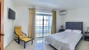 Una habitación de hotel con cama, sillón, televisión, mesita de noche y ventana con cortinas.