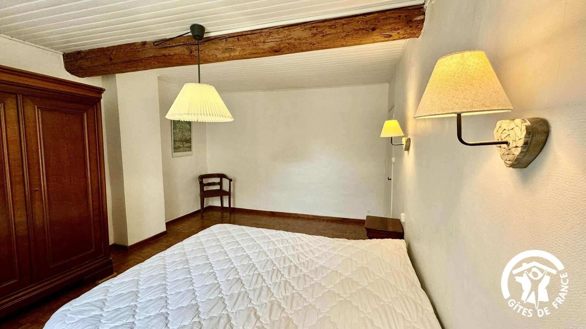 Une chambre avec un grand lit, des poutres apparentes, une armoire en bois, deux lampes fixées au mur et un tableau encadré accroché au mur.