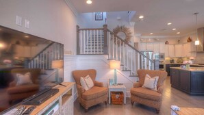 A spacious living room with a staircase leading to a kitchen area. There are two armchairs and a small table with a lamp. - 2 Homes, Sleeps 25 Pool, Firepit, Free Golf Carts Blue Mountain Beach (Santa Rosa Beach)