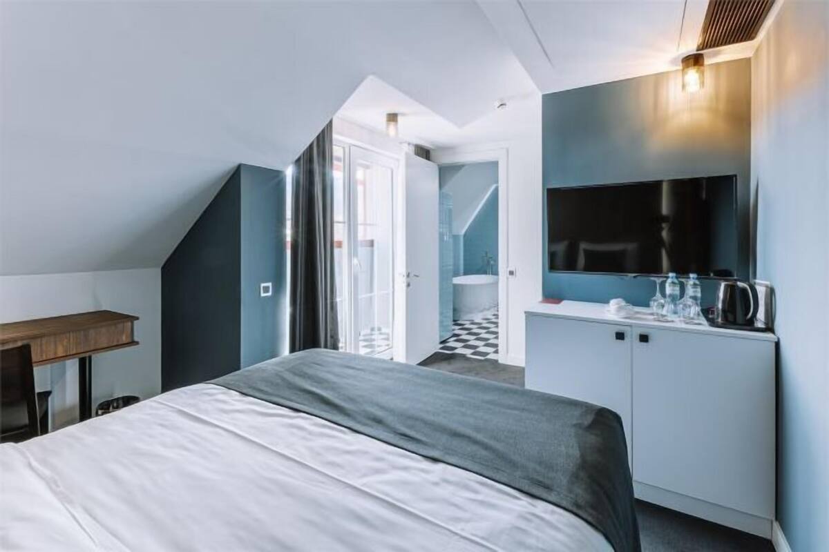 A bedroom with a bed, a desk, a mirror, and a kitchenette.