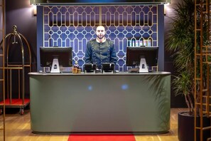 A reception desk with a modern design, two computer monitors, a small table with a bottle, and a decorative wall pattern. - Hotel Twenties Tbilisi - Stay & Dine (Tbilisi)