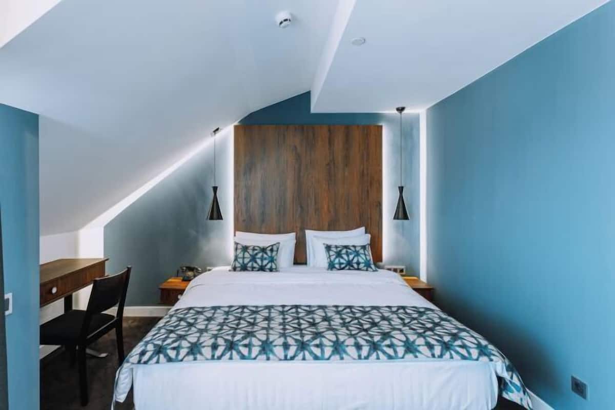 Ein Schlafzimmer mit schräg stehender Decke, einem Bett mit geometrischem Muster auf der Bettdecke, zwei Nachttischlampen und einem Holzkopfende.