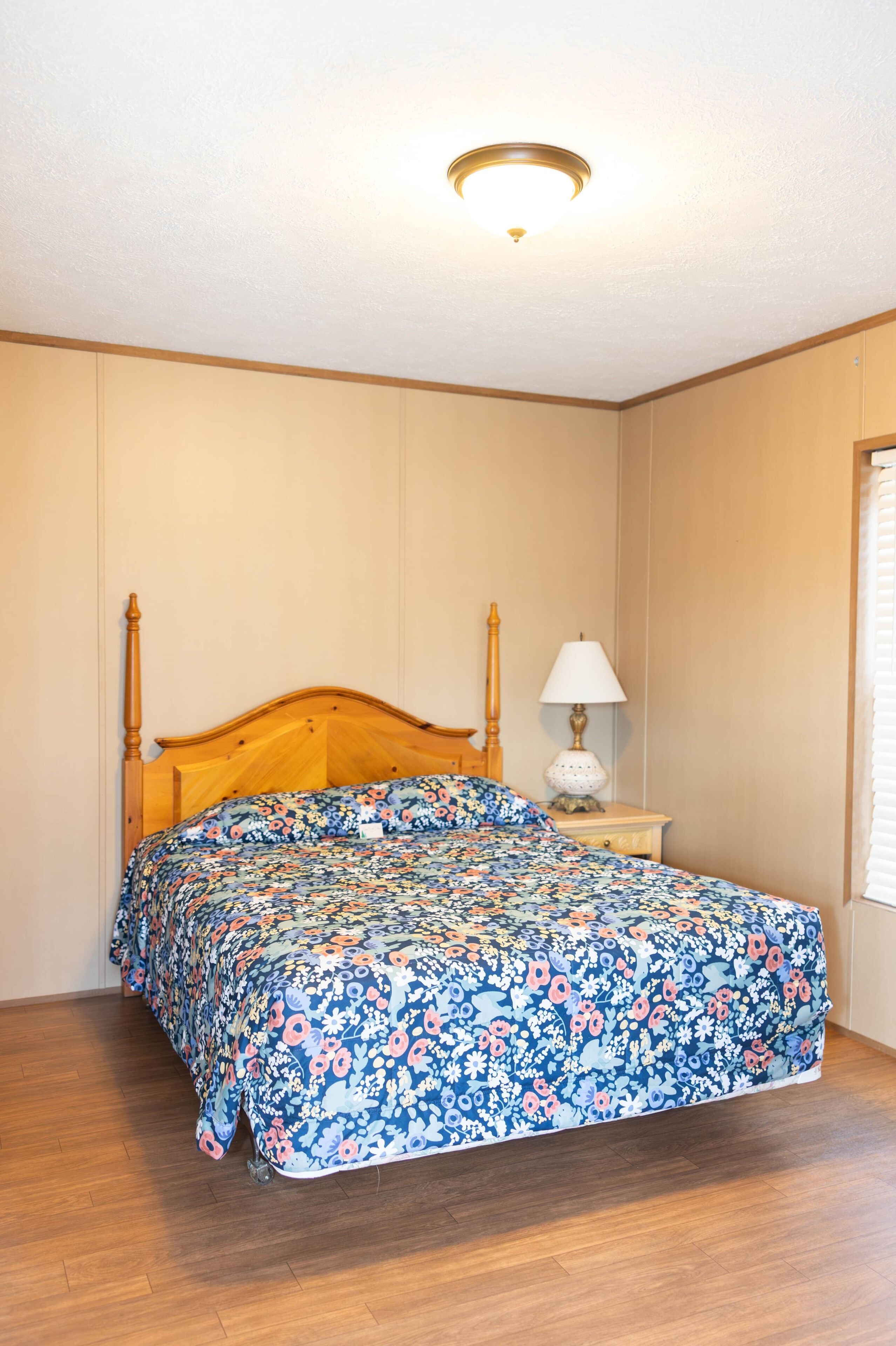A bedroom with a wooden bed, a nightstand with a lamp, and a window with blinds.