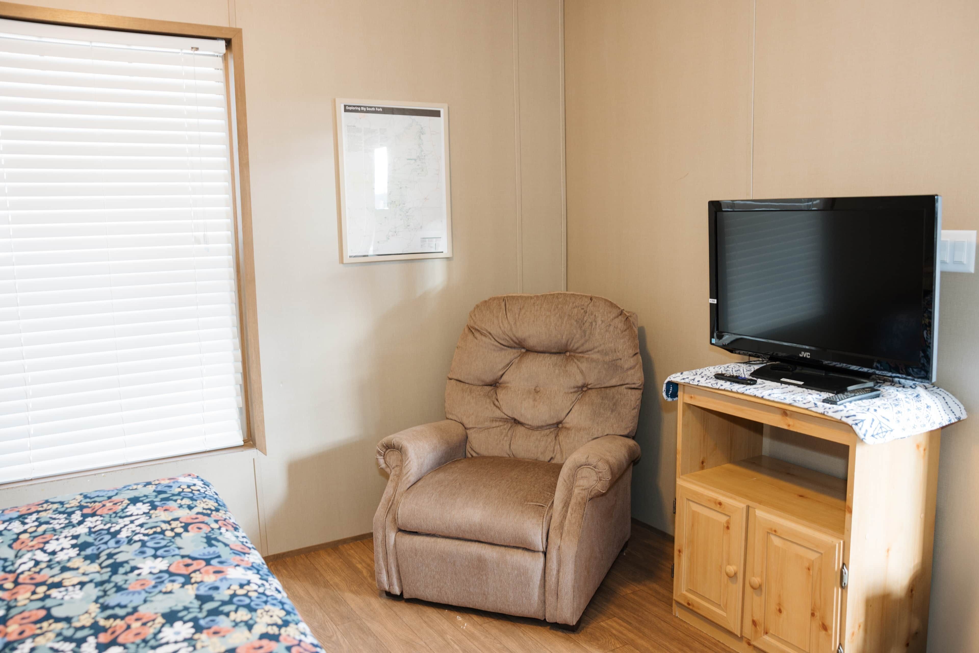 A room with a single bed, a cushioned armchair, a television on a wooden stand, and a map on the wall.