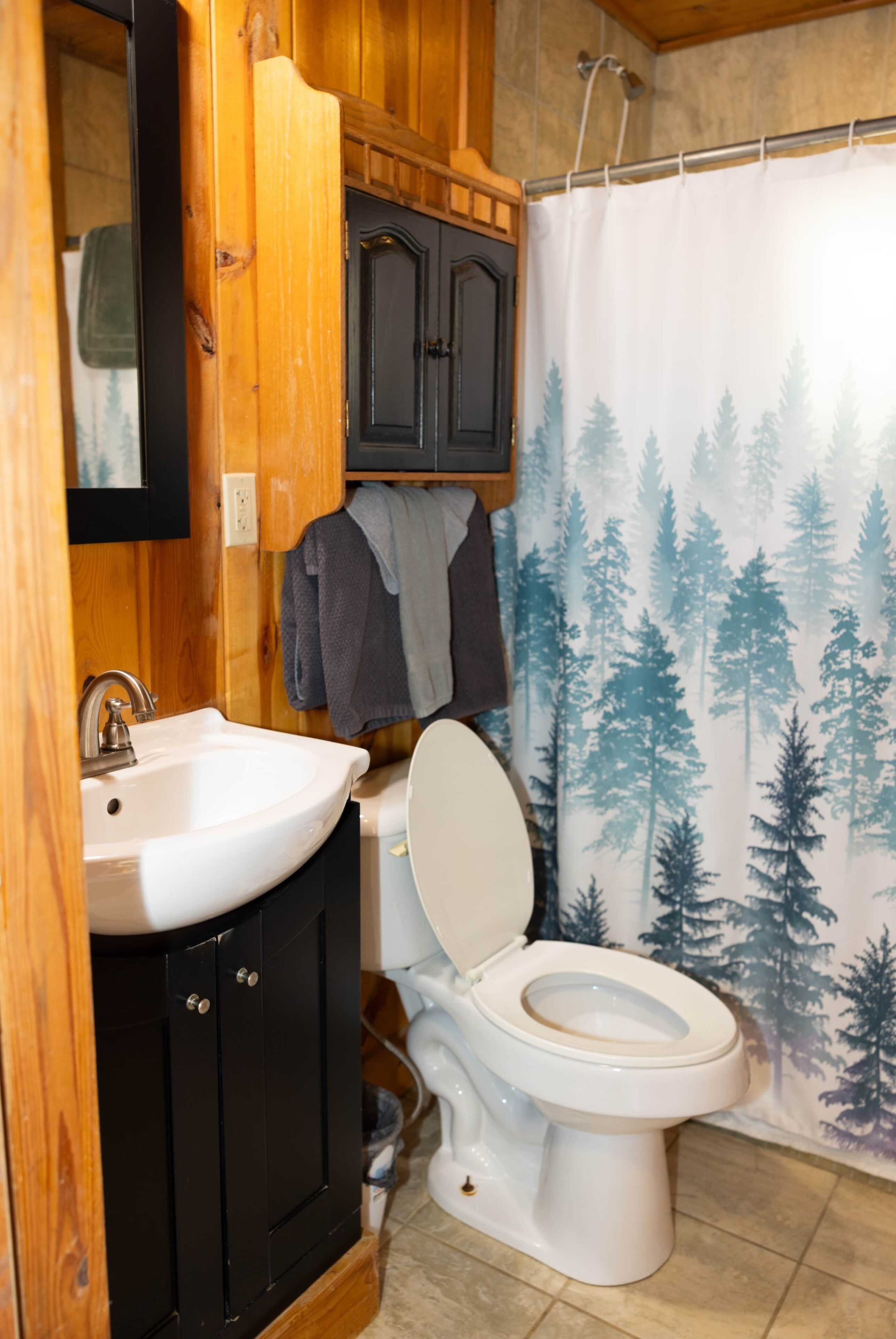 A bathroom with a white sink, a black cabinet, a toilet, and a shower curtain with a forest pattern.