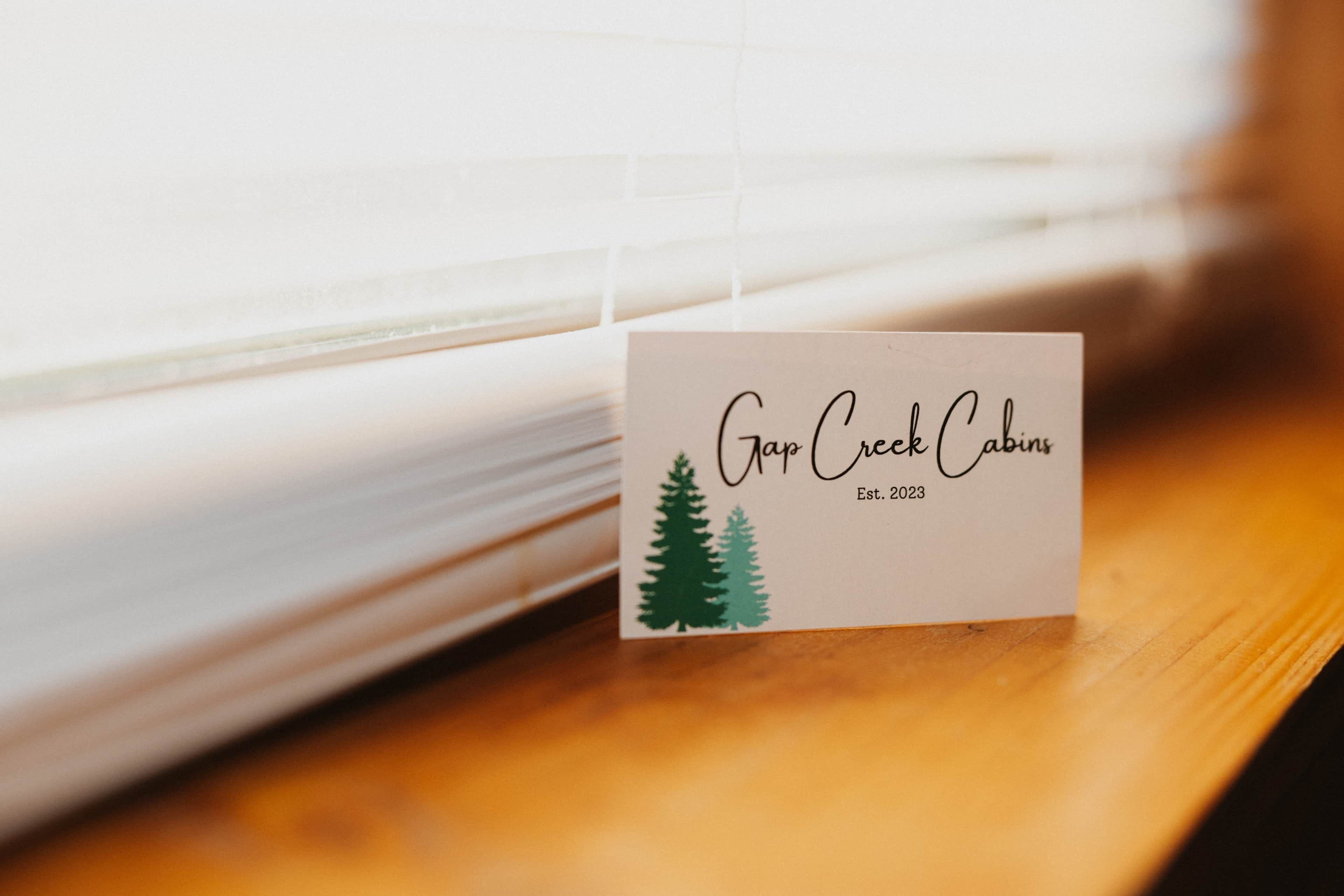 A business card for Gan Creek Cabins with a logo of two pine trees and the establishment year 2023.