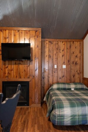 Ein Zimmer mit Holzwandverkleidung, einem flachbildfernseher über einem Kamin, einer karierten Bettdecke und Holzboden.