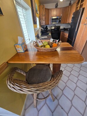 A kitchen with wooden cabinets, a microwave, a refrigerator, a wicker chair, and a table with a basket of fruit.