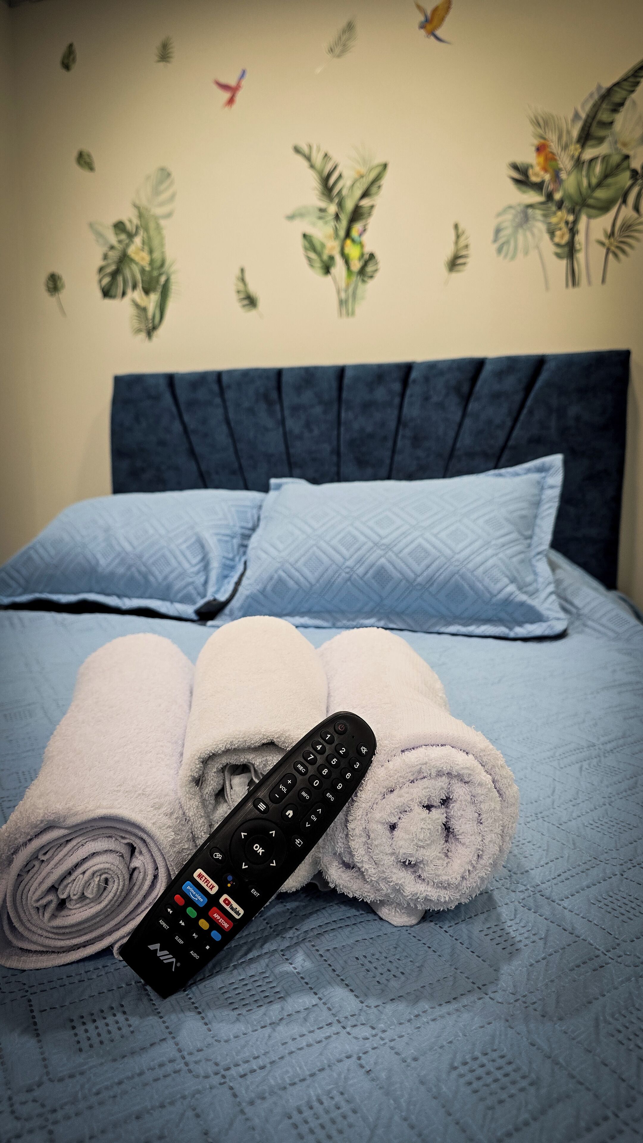 Ein ordentlich bezogenes Bett mit blauer Kopfteil und Kissen, ein aufgerolltes Handtuch und eine Fernbedienung liegen darauf.
