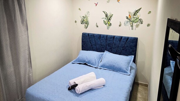 Un letto rifatto con cura, con copriletto e cuscini blu, una testiera blu scuro e un comodino con un asciugamano bianco e telecomando.