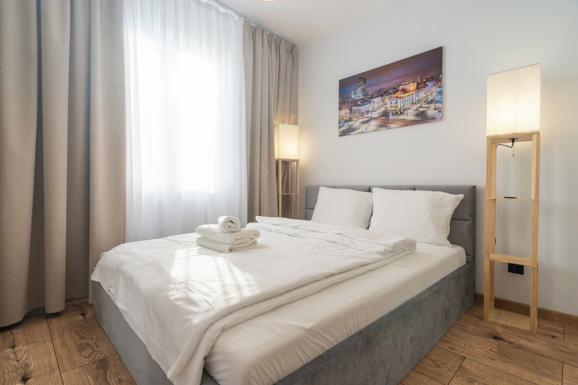 A hotel room with a large bed, a painting of a cityscape, a floor lamp, and a bedside table.