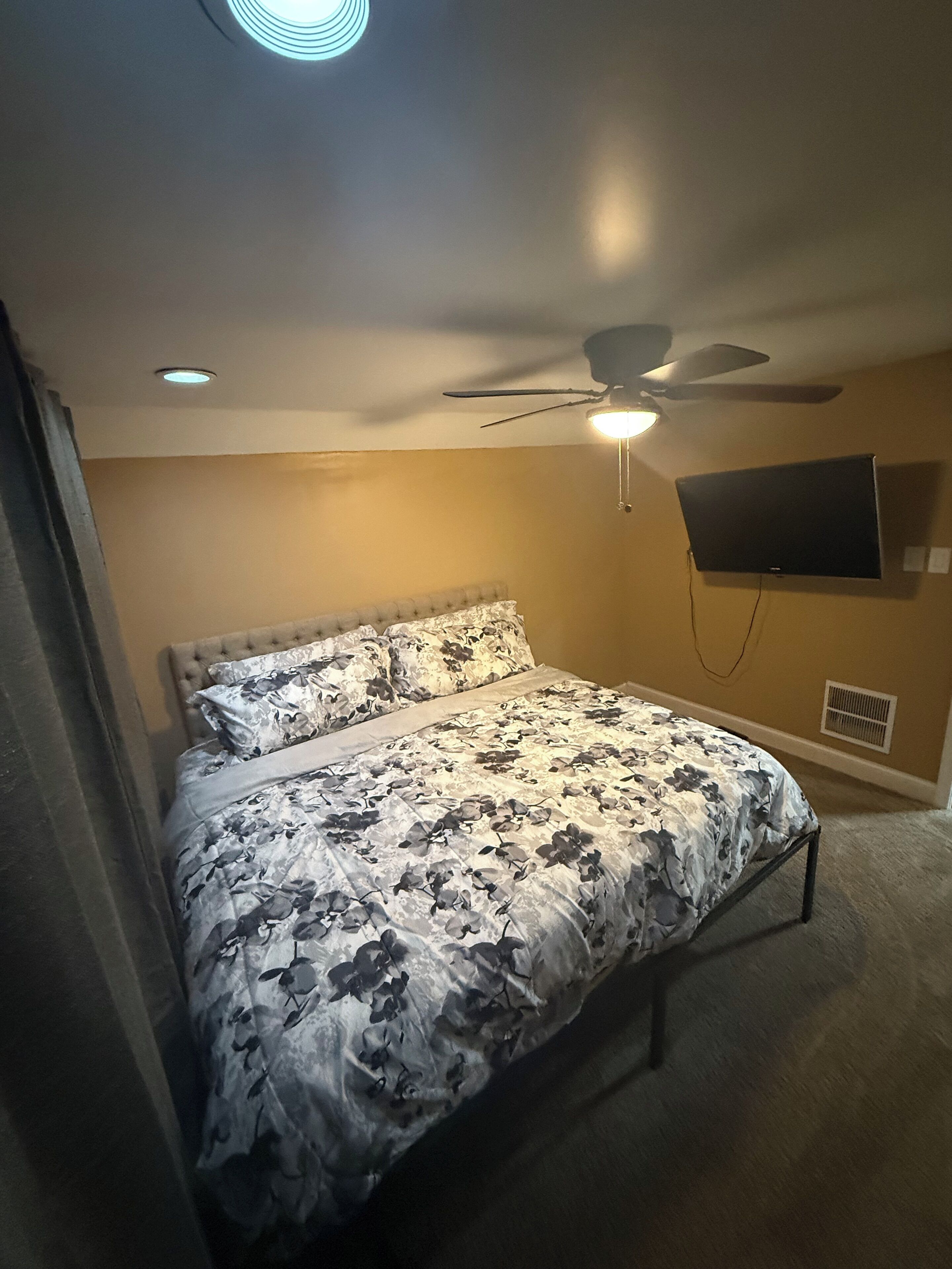 Un dormitorio con una cama, ventilador de techo, televisor montado y una rejilla de ventilación.