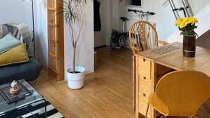 Ett kompakt bostadsutrymme med en säng på en loftvåning, en matplats, ett litet bord med stolar, en krukväxt och en cykel i bakgrunden.