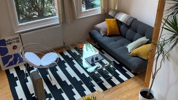 Ett modernt vardagsrum med en soffa, ett matbord och ett soffbord. Det finns stora fönster med gardiner, en hängande lampa och en krukväxt.