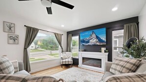 A modern living room with a fireplace, a large flat-screen TV, and a view of a green landscape. - Family Retreat Near Trails & Downtown • BBQ, Fire Pit & Kids’ Games (Ogden)