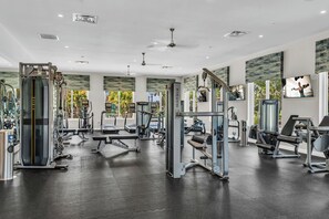 Ein Fitnessstudio mit verschiedenen Trainingsgeräten, einem Deckenventilator und großen Fenstern mit Blick auf Palmen.