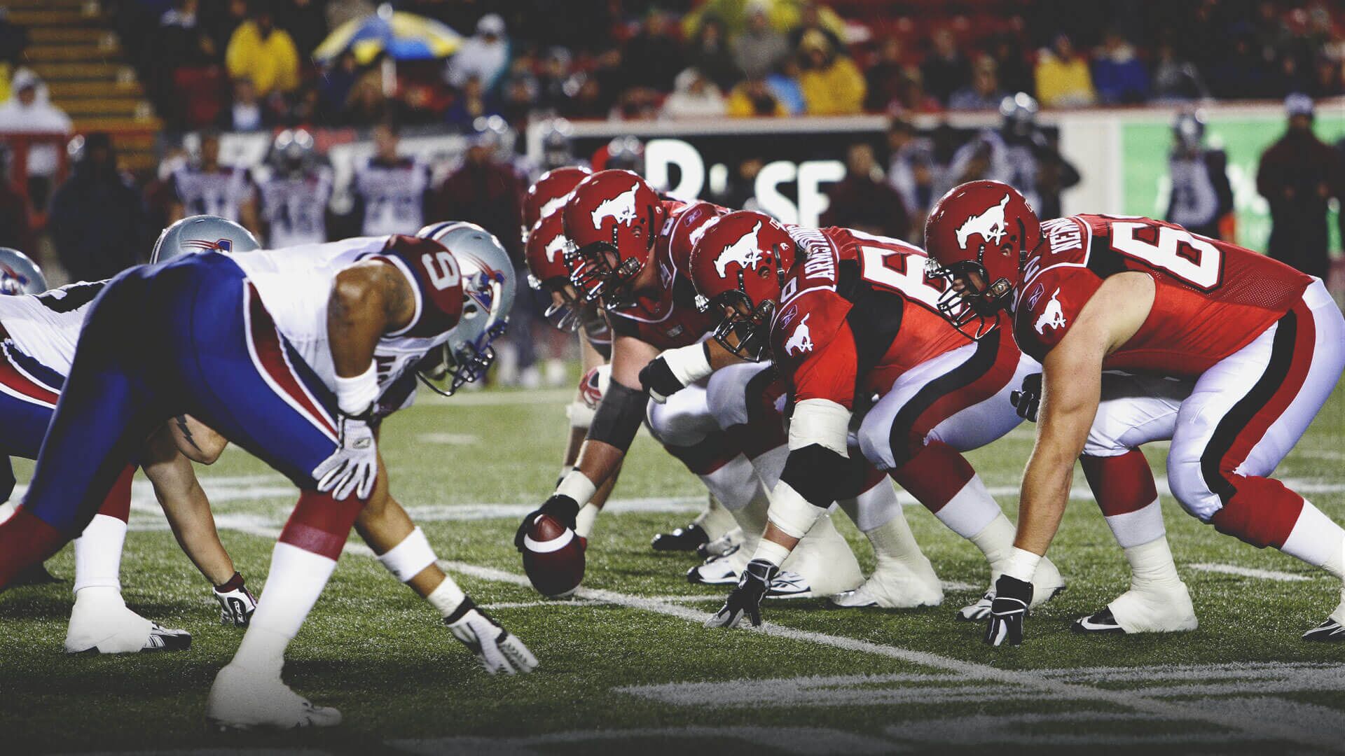 A football game with players in red and blue uniforms lined up at the line of scrimmage.