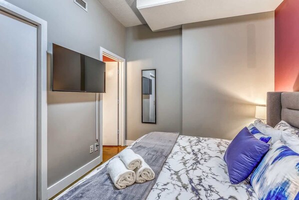A modern bedroom with a large bed, a flat-screen TV mounted on the wall, and a mirror reflecting the room. - Stylish Condo With Balcony in Downtown Calgary (Calgary)