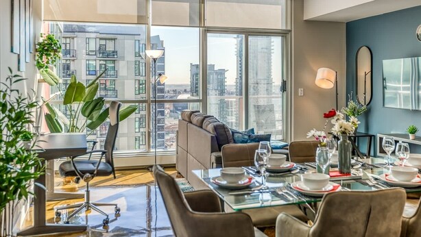 Una sala de estar moderna con un amplio ventanal corredera que da a un balcón, un sofá, una mesa de comedor preparada para cuatro personas y vistas a la ciudad.