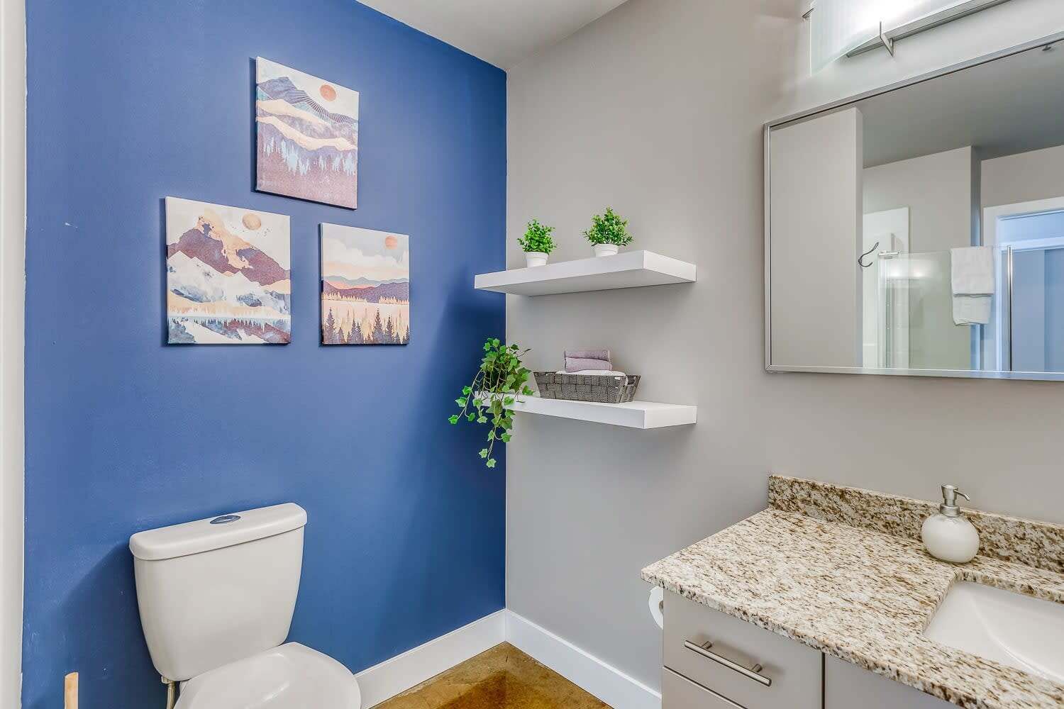 Un baño con pared azul, inodoro blanco, encimera de granito y espejo sobre el lavabo.