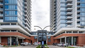 A modern building with multiple floors, large windows, and a central entrance with a decorative light fixture. - Newly Built Modern Condo w Balcony & City Views (Kitchener)