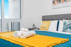 Una habitación de hotel moderna con un ventanal grande, una cama bien hecha con colcha amarilla y azul, y una almohada con el texto 'home sweet home'.