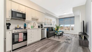A modern kitchen with white cabinetry, a built-in microwave, and a stainless steel oven. There is a gray sofa and a coffee table in the living area, and a large window with blinds. - Newly Built Modern Condo w Balcony & City Views (Kitchener)