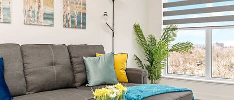 Ein modernes Wohnzimmer mit einem grauen Sofa, gelb-blauen Kissen, einem Glastisch mit Blumen und einem großen Fenster mit Blick auf Bäume.