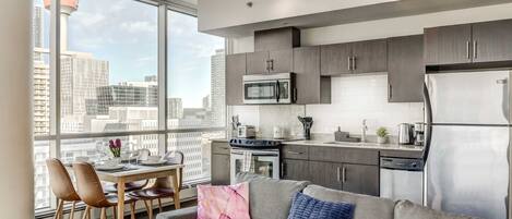 A modern kitchen with stainless steel appliances, a dining area with a table and chairs, and a grey sofa with decorative pillows.