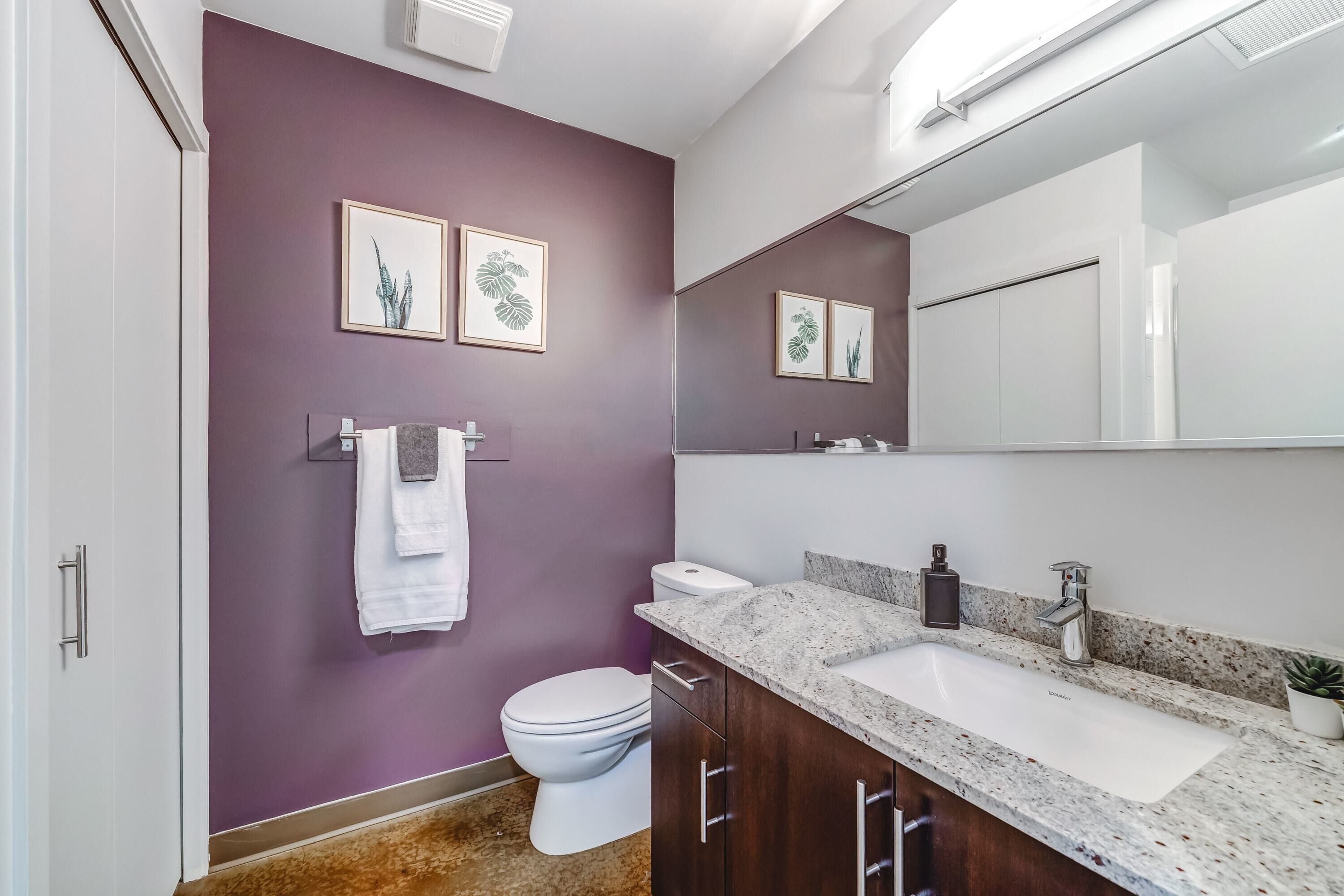 Une salle de bain moderne avec un mur violet, des toilettes blanches, un grand miroir et un lavabo sur un comptoir en granit.