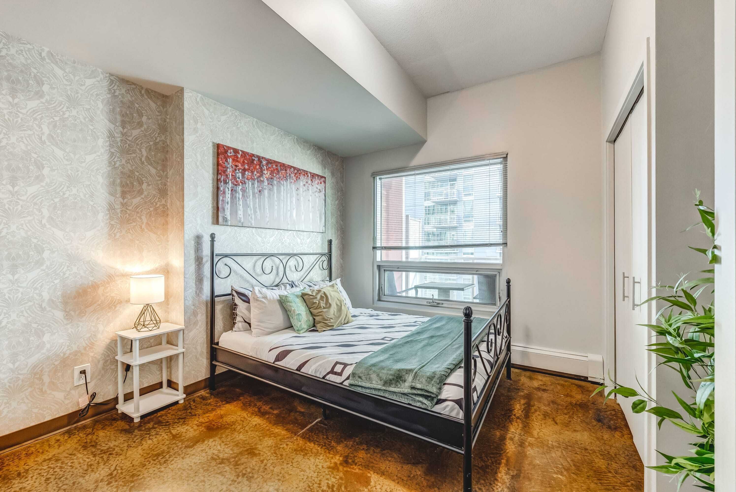 A bedroom with a bed, a nightstand, a window, and a plant.