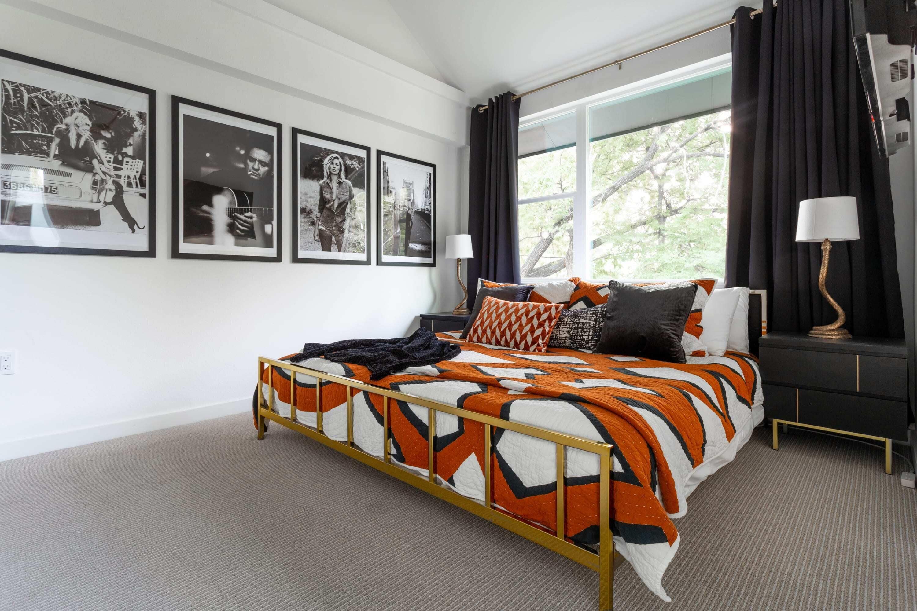 A bedroom with a bed, framed pictures on the wall, a window with curtains, and a nightstand with a lamp.