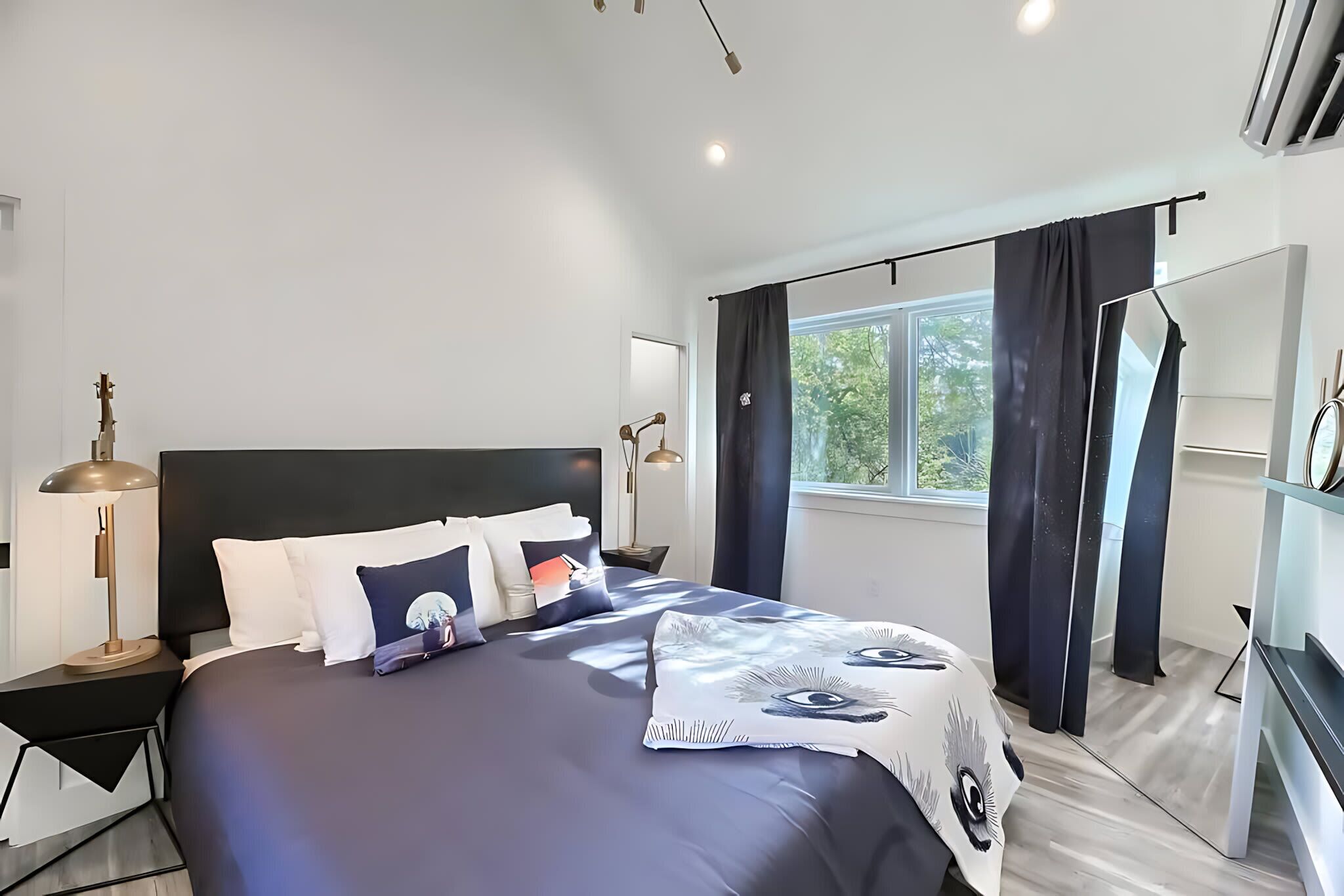 A modern bedroom with a large bed, a nightstand with a lamp, a dresser, and a window with curtains.