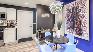 A modern kitchen with white cabinets, a microwave, and a stove. There is a dining area with a black table, blue chairs, and a neon sign. - Stylish 3BR East Side Retreat w Patio & BBQ (Austin)