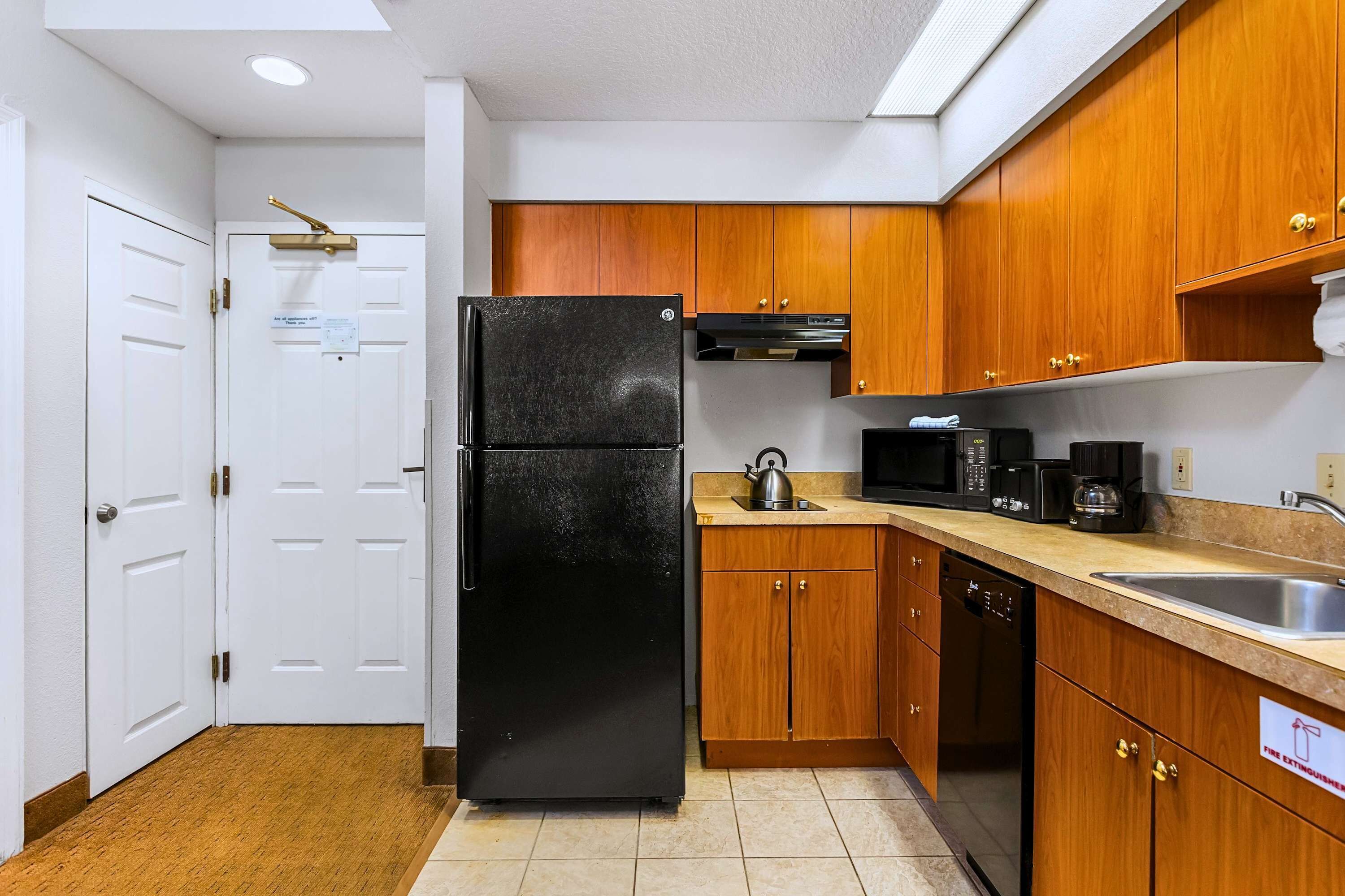 A compact kitchen with wooden cabinets, a black refrigerator, a microwave, a kettle, and a coffee maker.