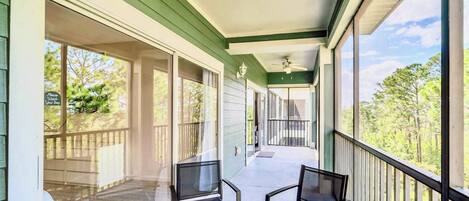 Un balcon abrité, équipé de deux chaises, d’un ventilateur de plafond et offrant une vue sur les arbres et une terrasse en bois.