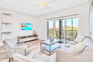 Un salon moderne avec un canapé, des fauteuils, une table basse et un téléviseur. Il y a un balcon avec vue sur les arbres et une rambarde.
